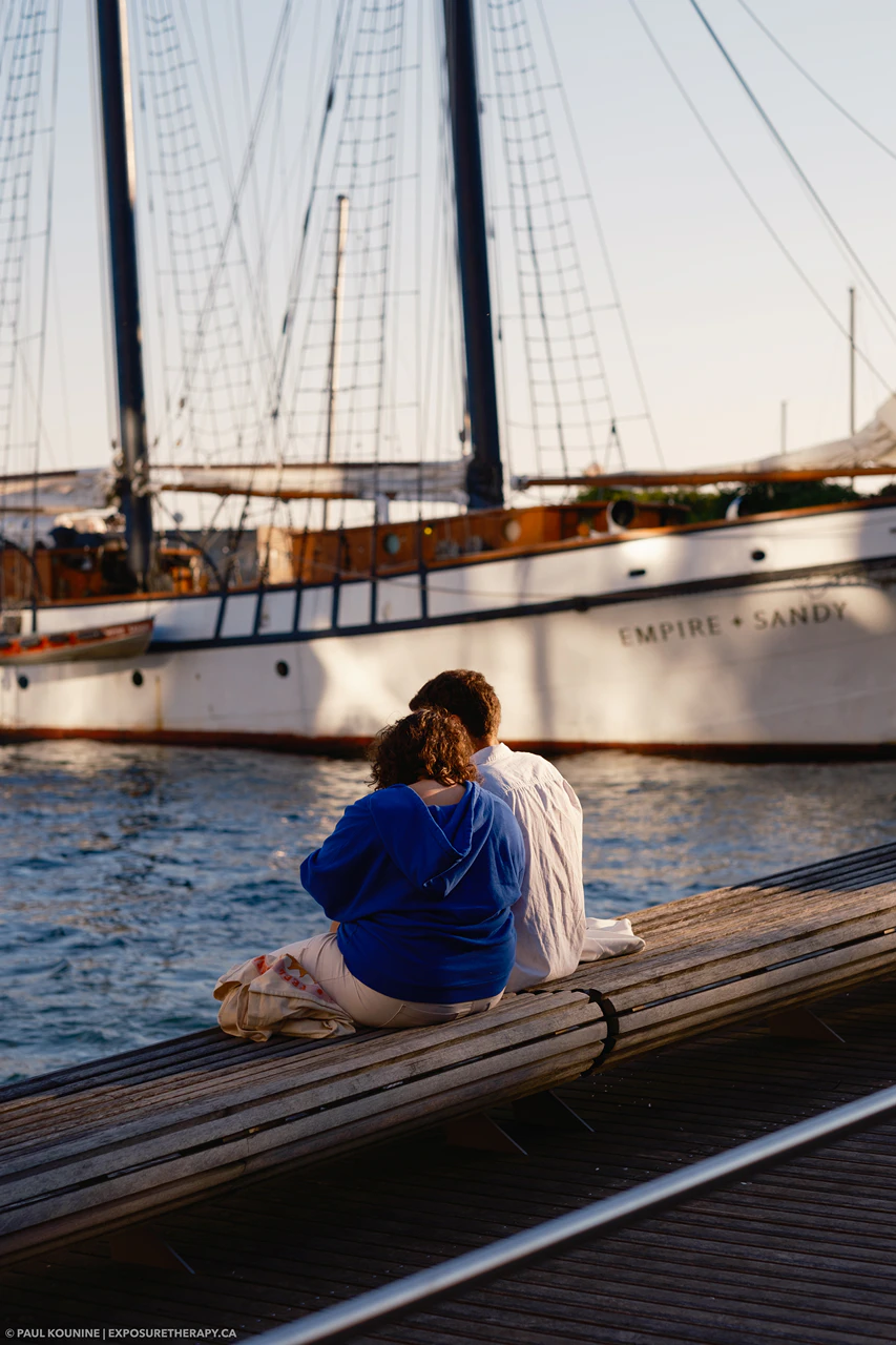 Couple sitting on wave deck on the Toronto waterfront with a large sailboat in background.